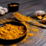 Pó de cúrcuma vibrante em uma tigela preta e em uma colher de metal sobre uma mesa de madeira escura, acompanhado de raízes de cúrcuma frescas ao fundo.