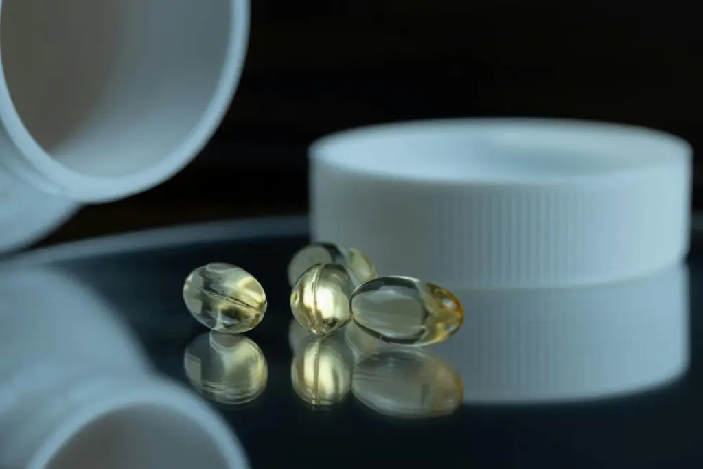 Cápsulas de ômega-3 douradas e translúcidas sobre uma superfície reflexiva ao lado de um frasco branco, destacando a pureza e o aspecto límpido do óleo de peixe.