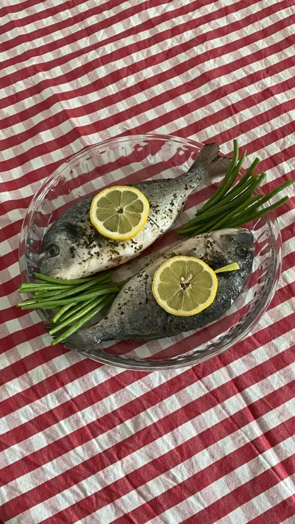 Dois peixes inteiros temperados com rodelas de limão e ervas em um prato sobre toalha xadrez vermelha.